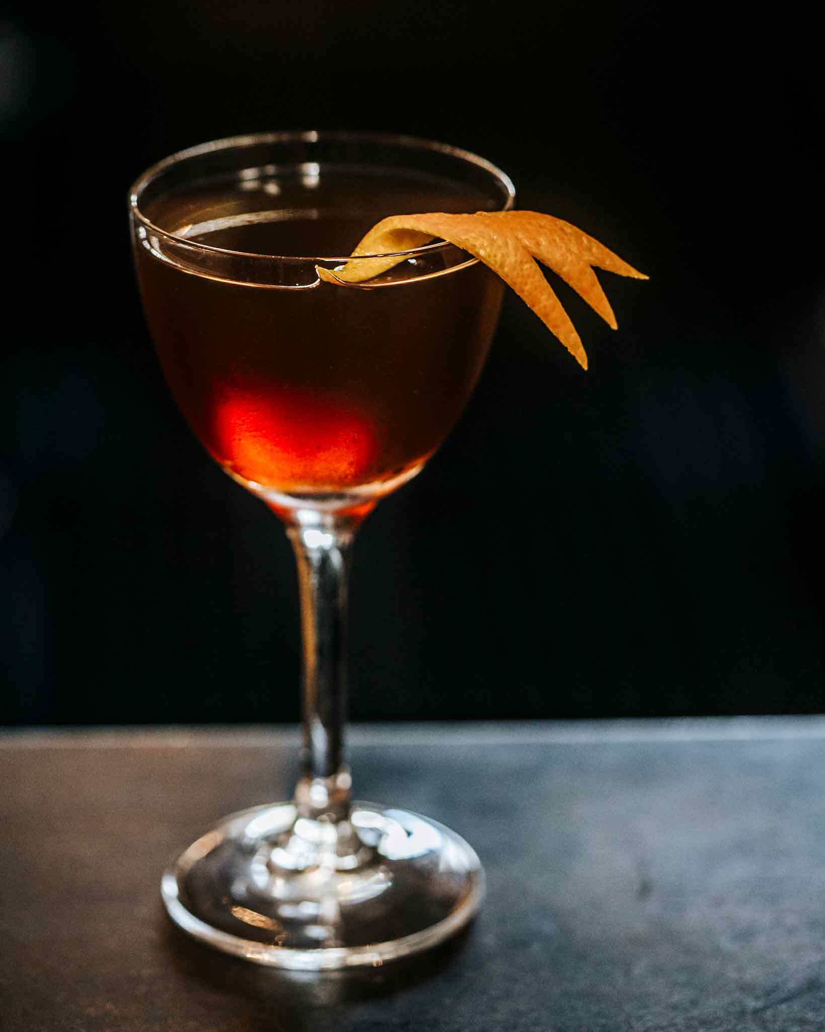 Bijou cocktail on bar in front of dark background with orange twist