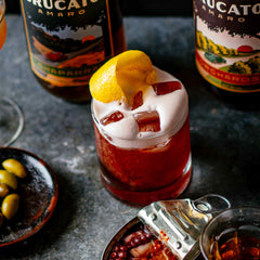 Fiddler's Green cocktail with orange twist in front of Brucato Amaro Bottles