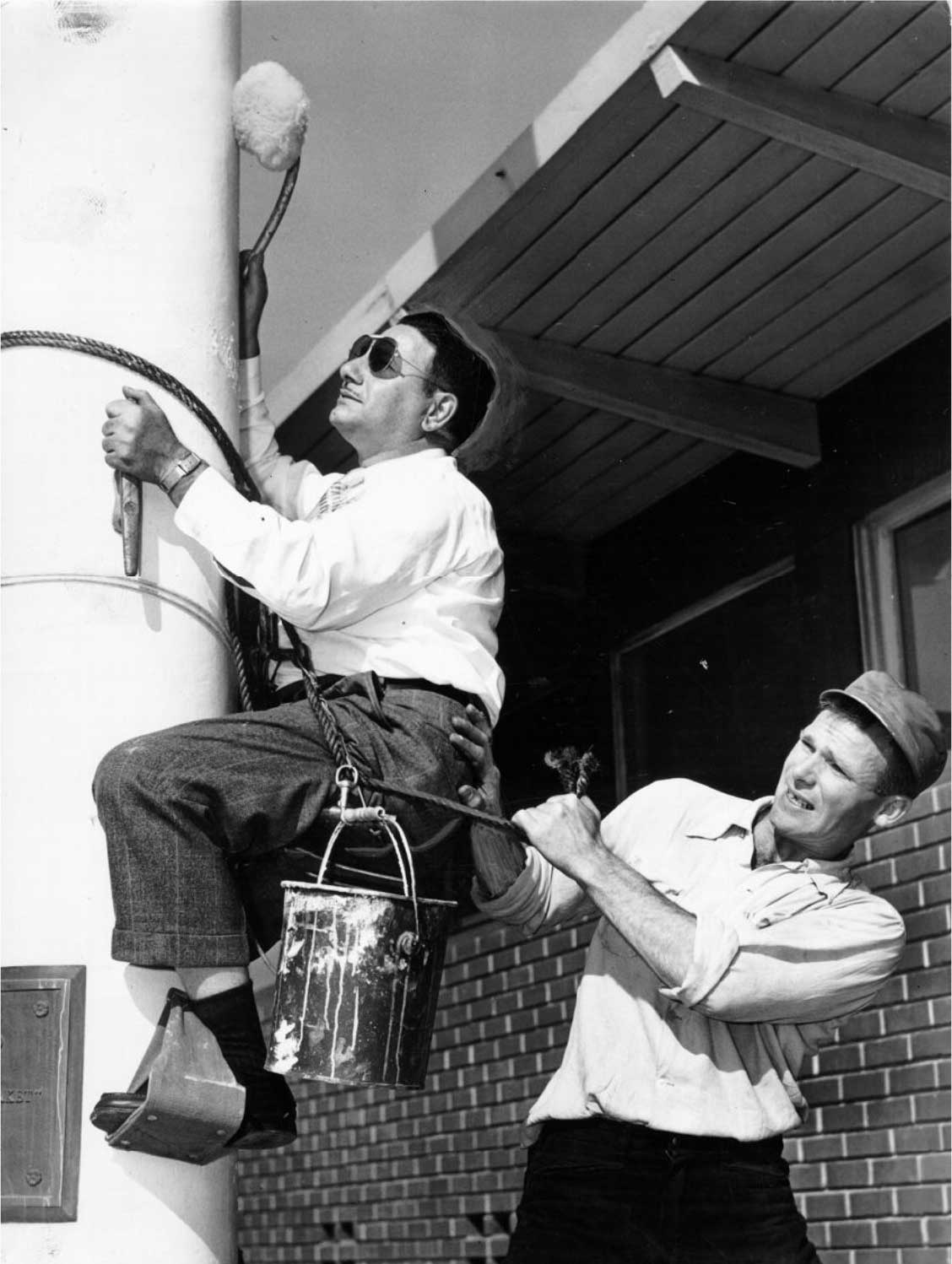 John Brucato painting a pole while man supports him from rope